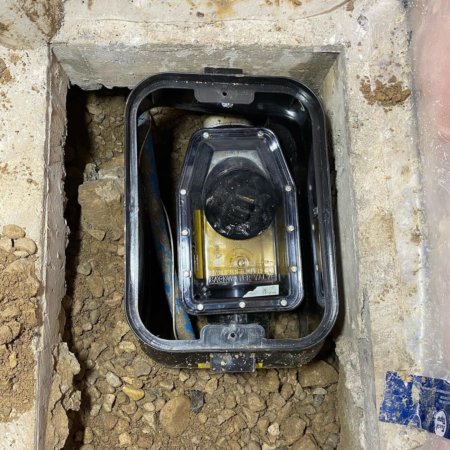 A water meter housed in a black rectangular plastic box set into a hole in the ground filled with loose soil and rocks.