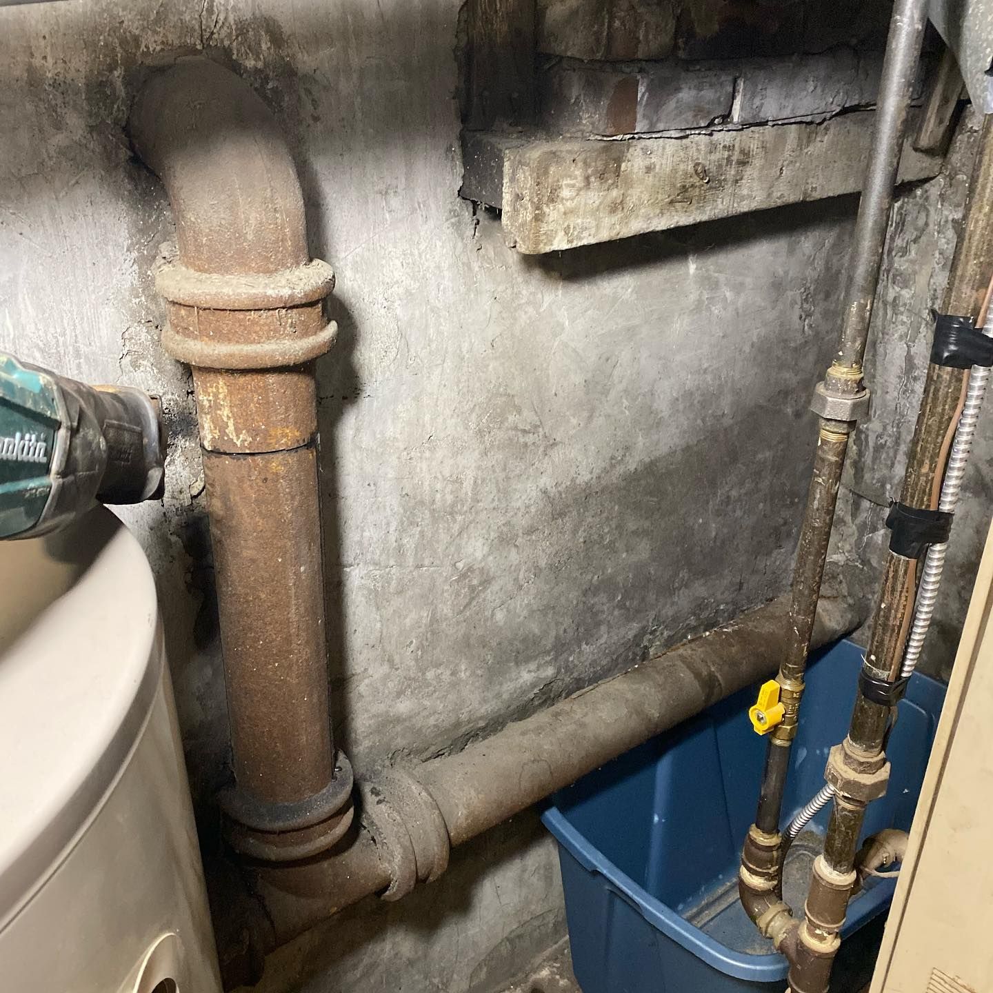 Cast iron pipes and a water heater in a basement with a blue plastic bin underneath the plumbing.
