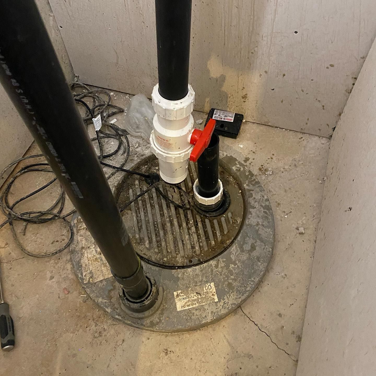 A sump pump basin in a basement floor with black piping, a white check valve, and a red ball valve handle.