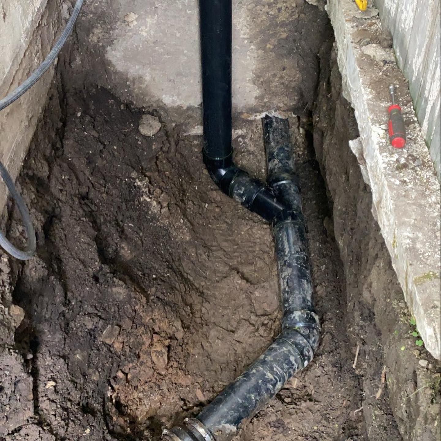 A black plastic drainage pipe connects to a main horizontal pipe in a narrow, dirt-filled trench.