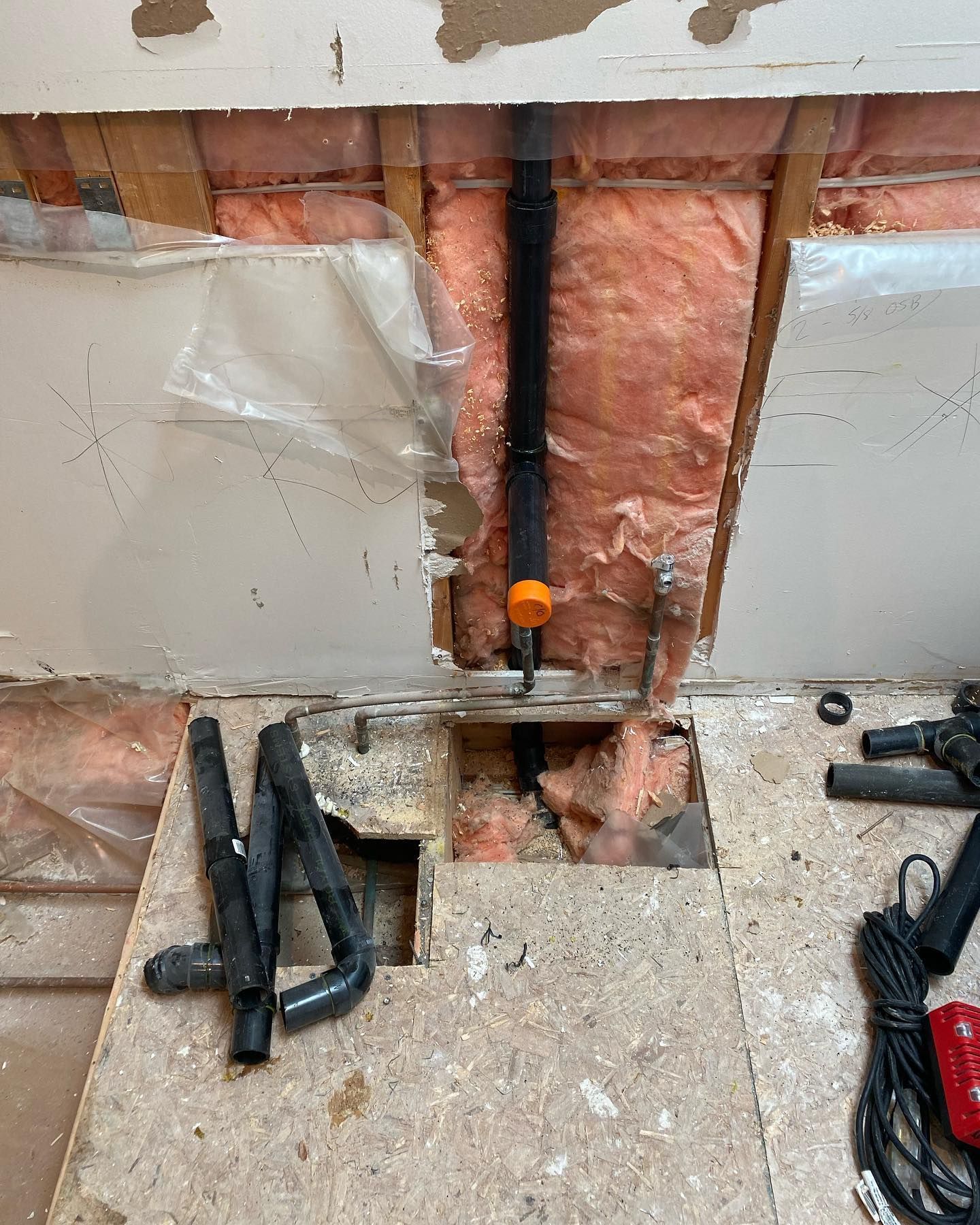A wall interior with pink insulation, a vertical black drain pipe with an orange cap, and exposed subflooring with pipes.