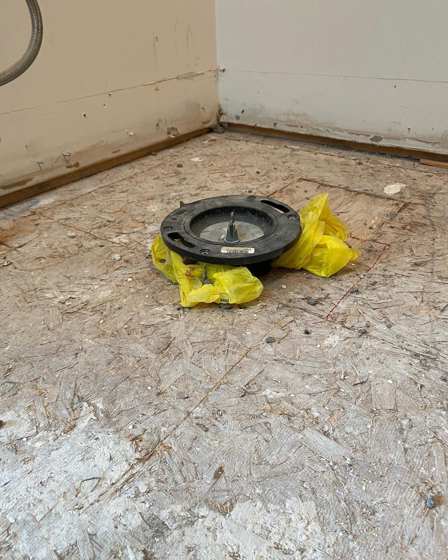 A black plastic toilet flange installed on a bare plywood subfloor, surrounded by crumpled yellow plastic.