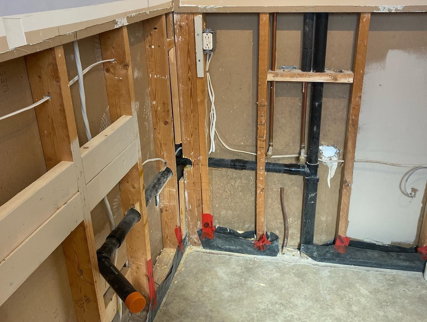 Exposed wooden wall studs in a room under construction, featuring visible electrical wiring and plumbing pipes.