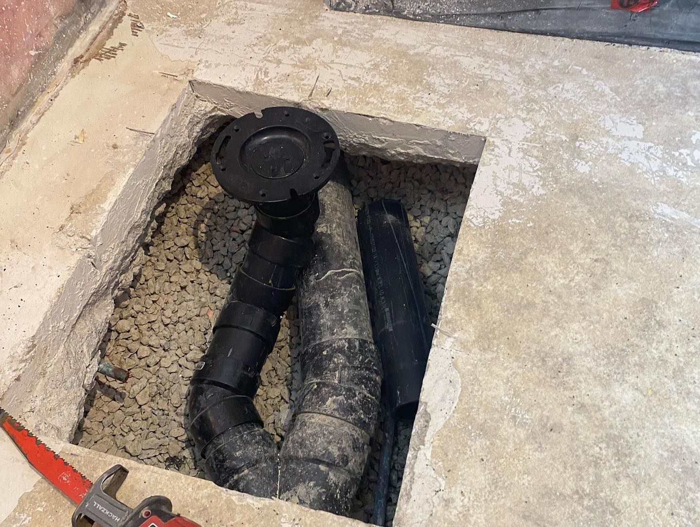 A black toilet flange and piping are set within a square hole cut into a concrete floor, with a tool visible nearby.