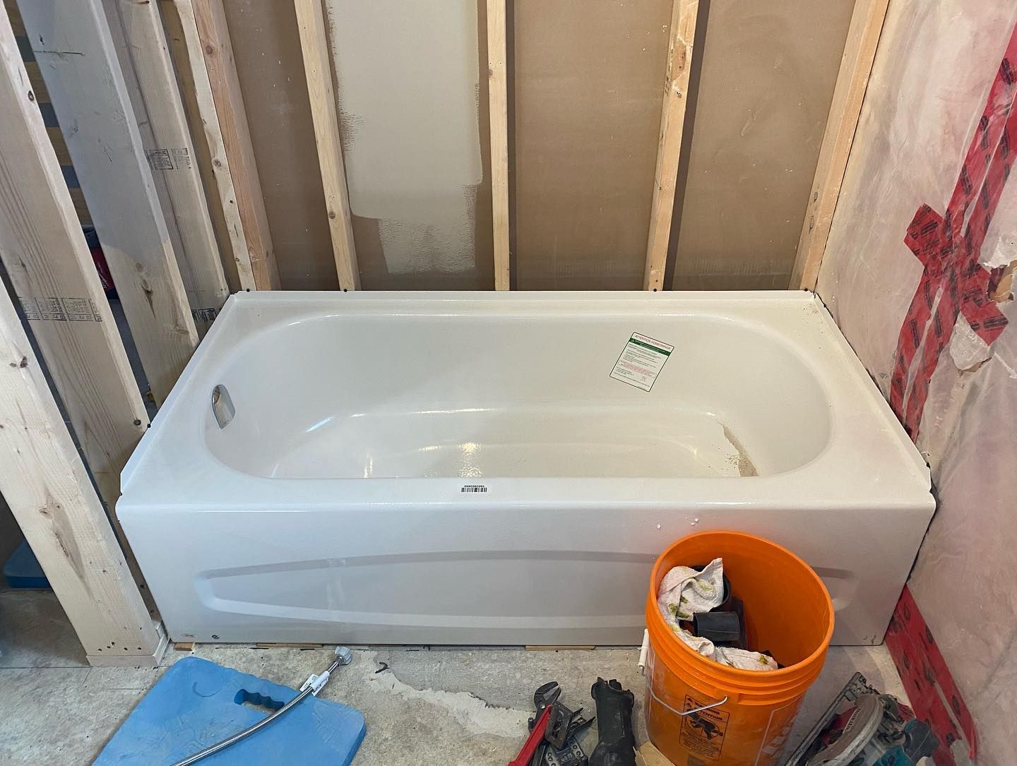 Bathtub installed in unfinished bathroom with exposed wall studs and an orange bucket nearby