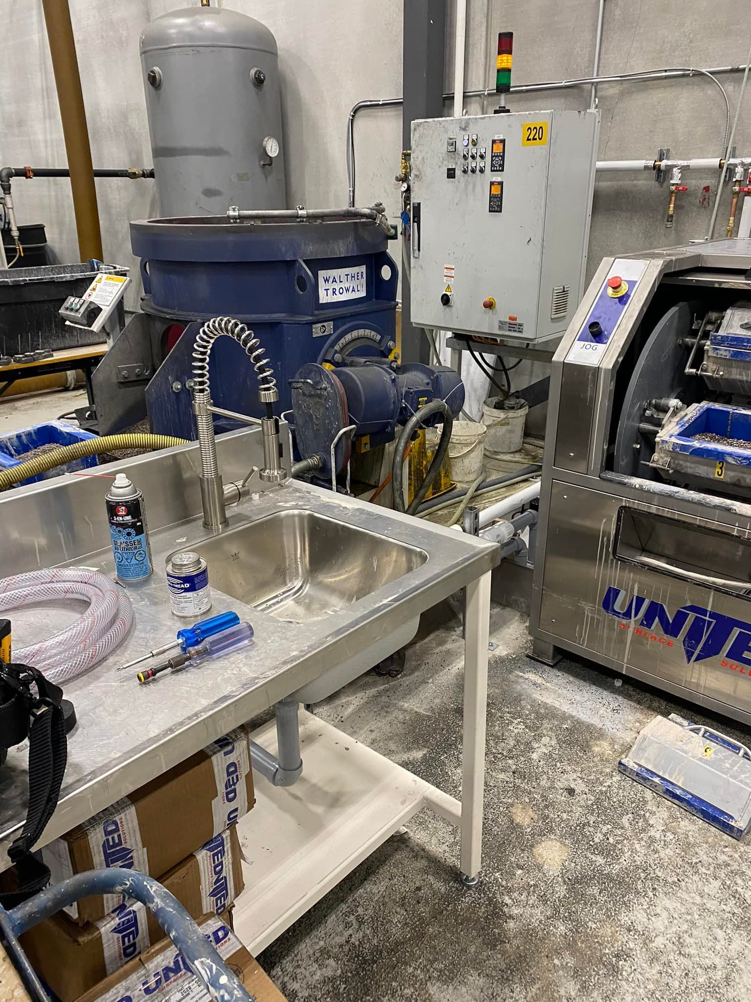 Industrial metal sink in a workshop area with industrial machinery, control panels, and a pressurized air tank behind it.
