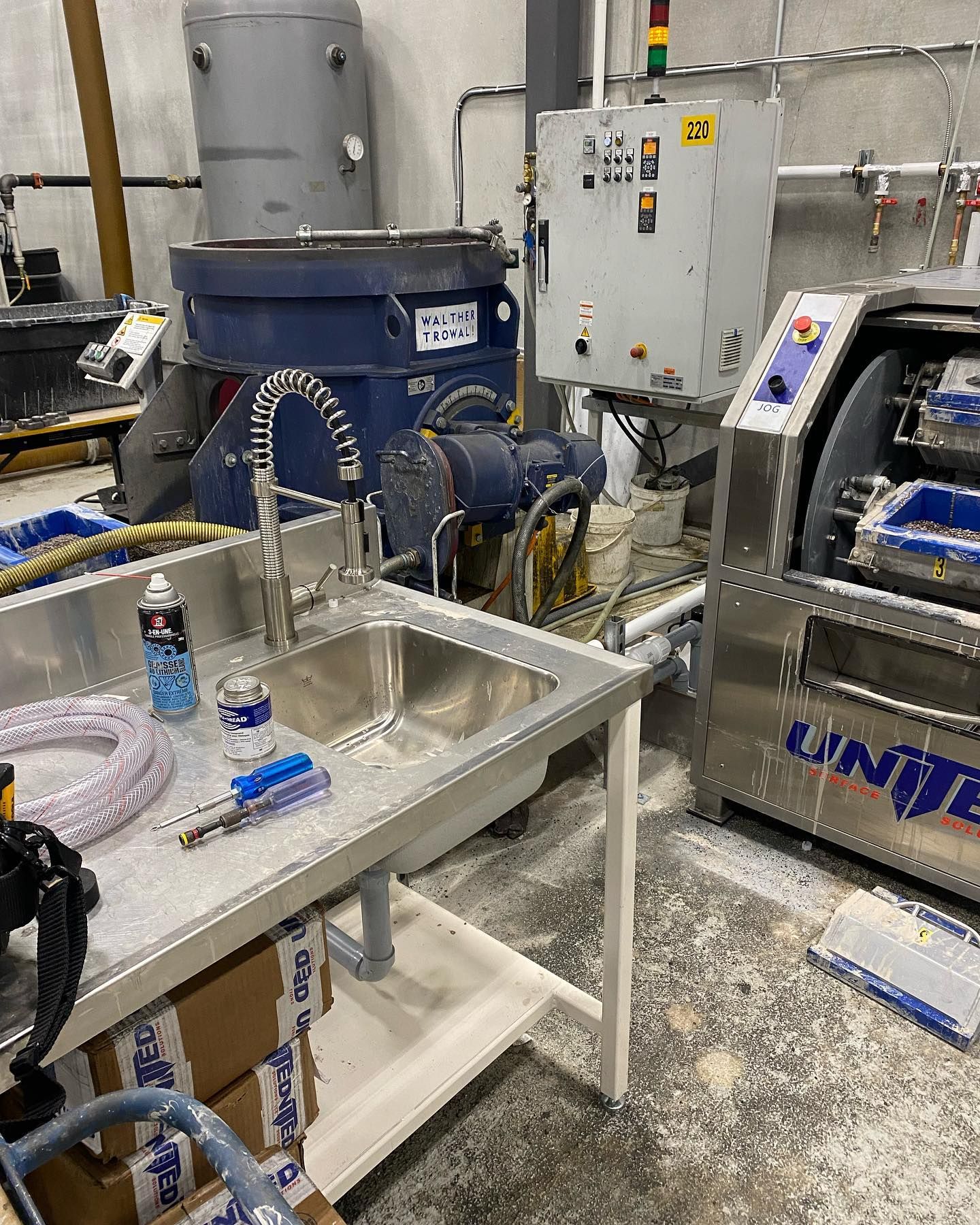 A stainless steel sink with a faucet sits in a workshop next to industrial machinery and a control panel.
