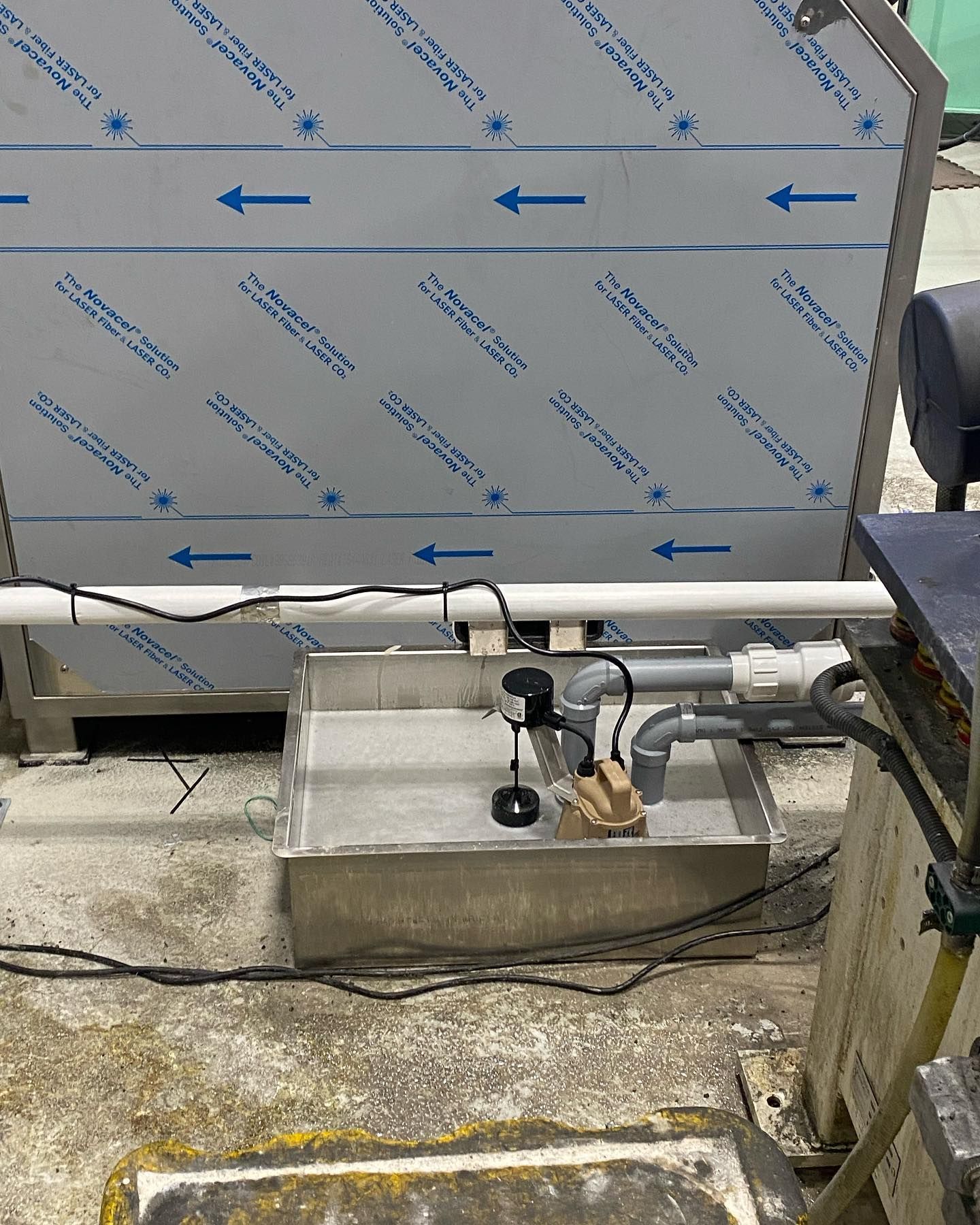 A stainless steel basin containing a black pump and grey PVC pipes sits on a concrete floor near industrial equipment.