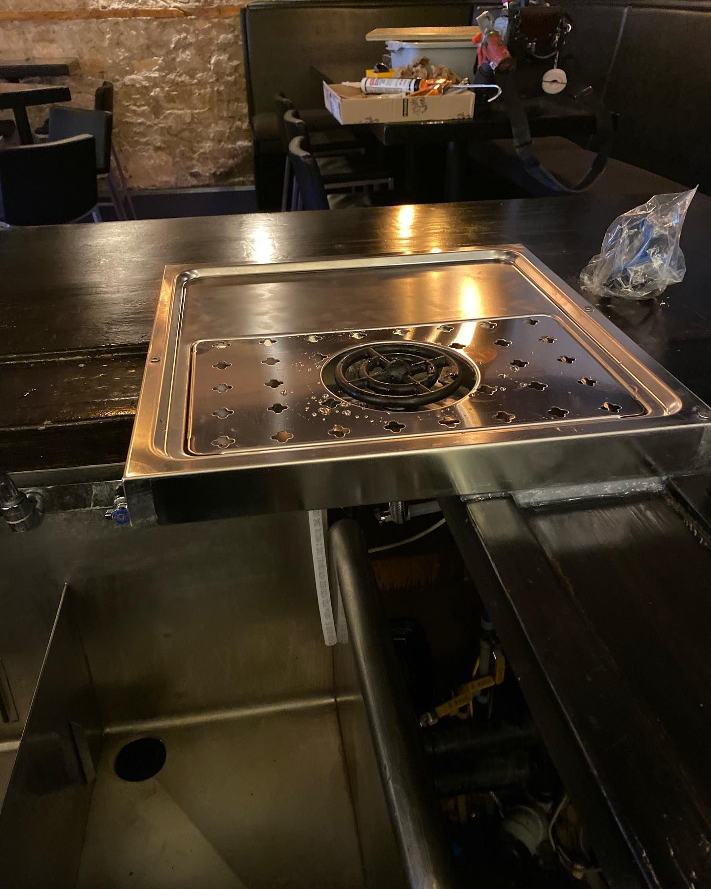 A metal bar drip tray with a circular drain sits on a dark wooden counter above a stainless steel sink.