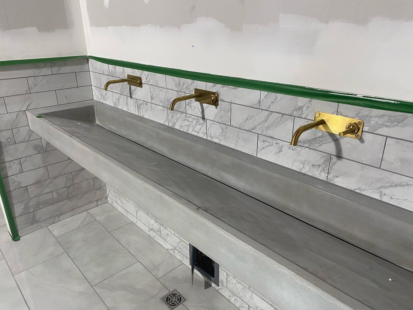 A long concrete trough sink with three brass wall-mounted faucets against a marble-tiled wall in a room under construction.