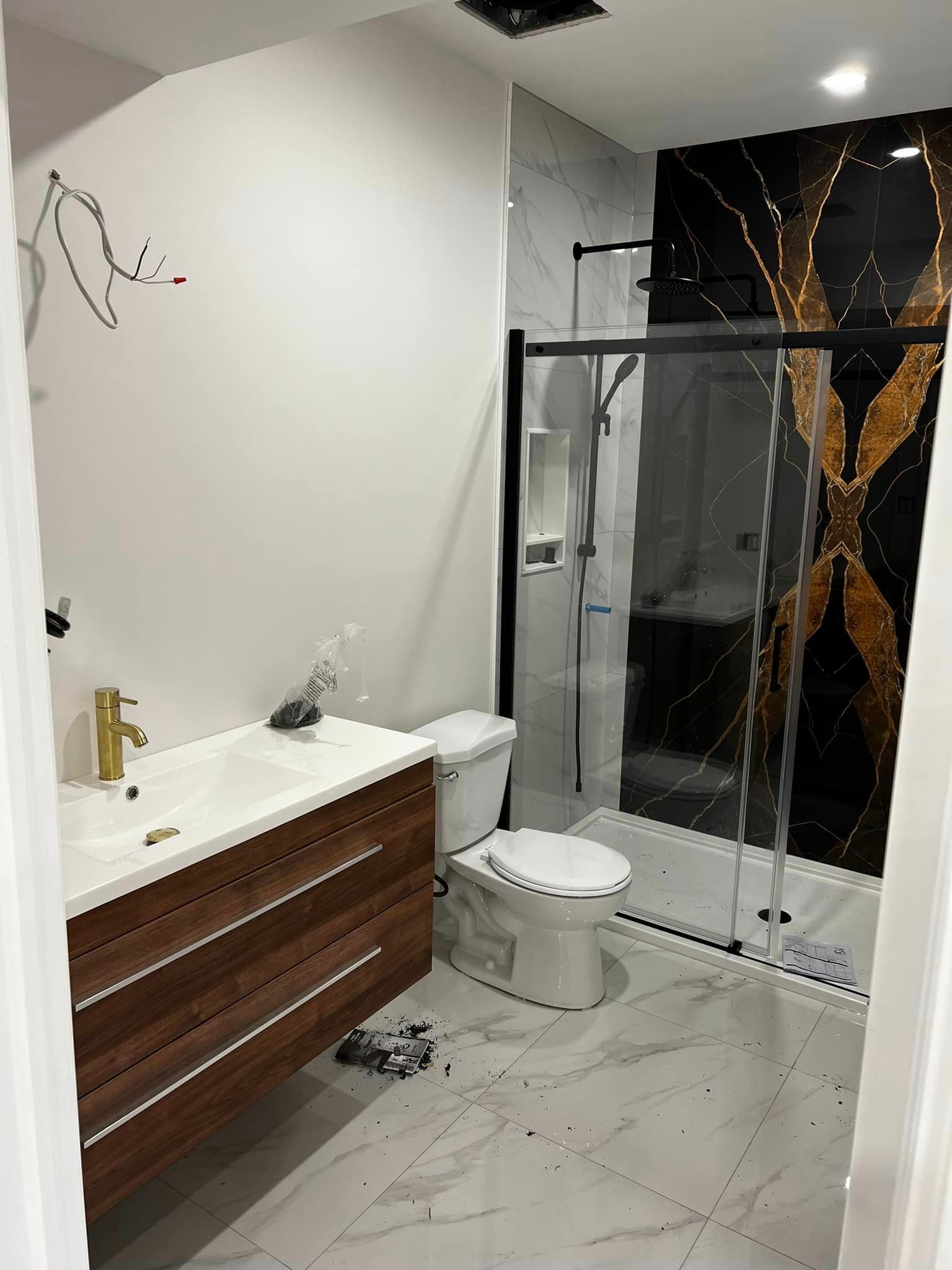 A modern bathroom with a wood vanity, white toilet, and a glass-enclosed shower featuring a bold, black and gold backdrop.