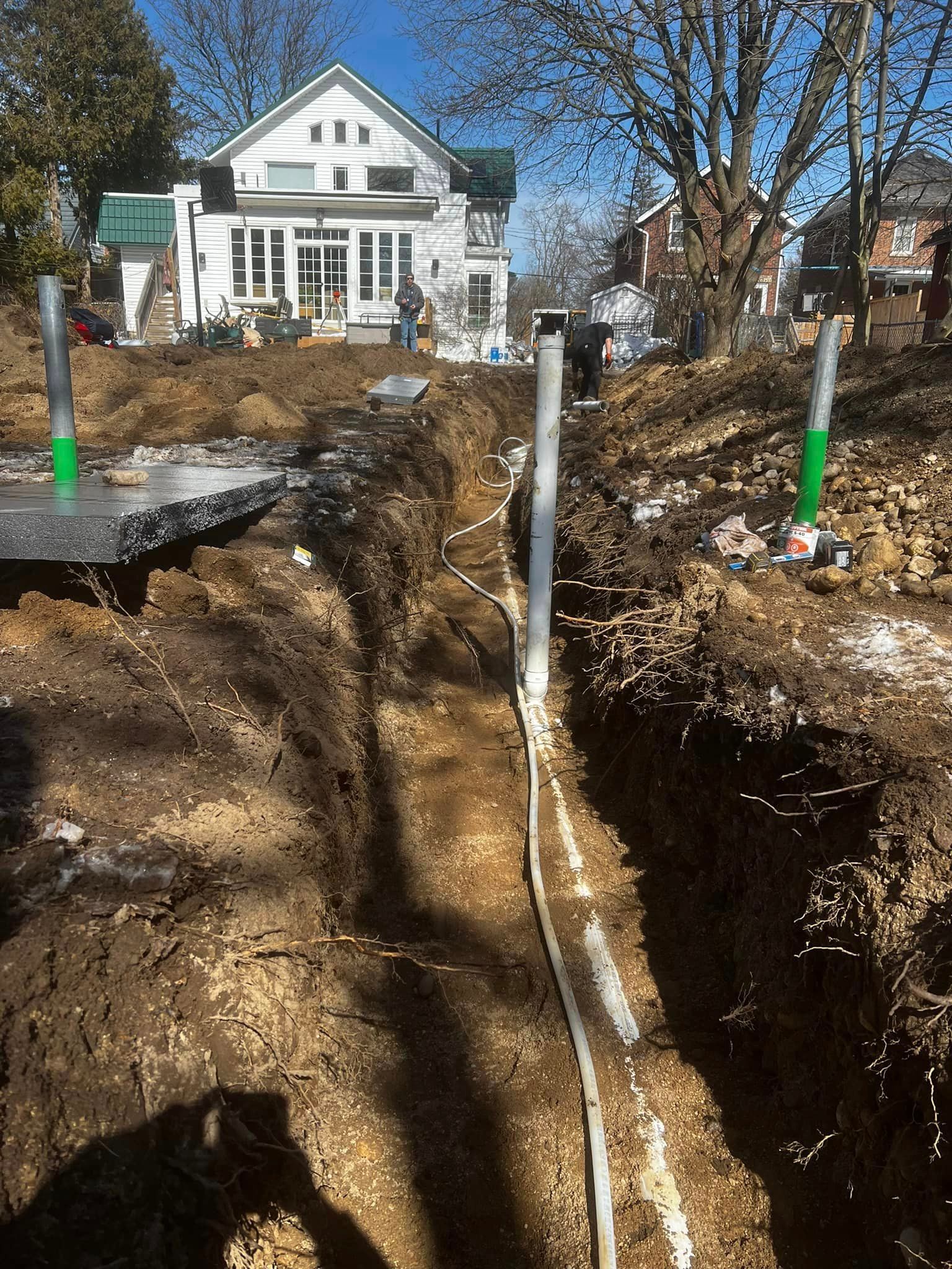 A residential construction site features a deep trench in the foreground with vertical utility posts in a grassy yard.