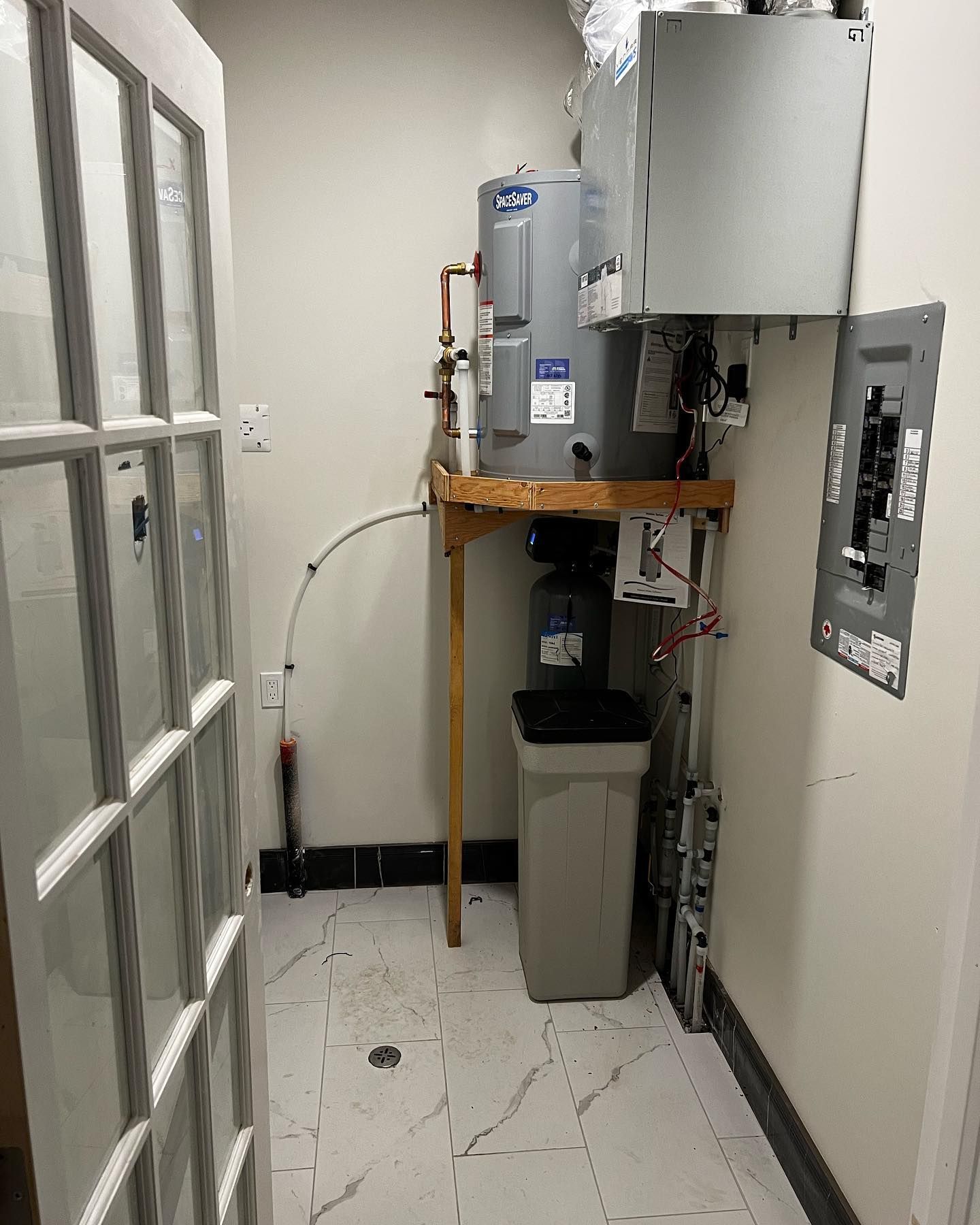 A small utility closet containing a water heater, a softener unit, and an electrical breaker panel on the white wall.