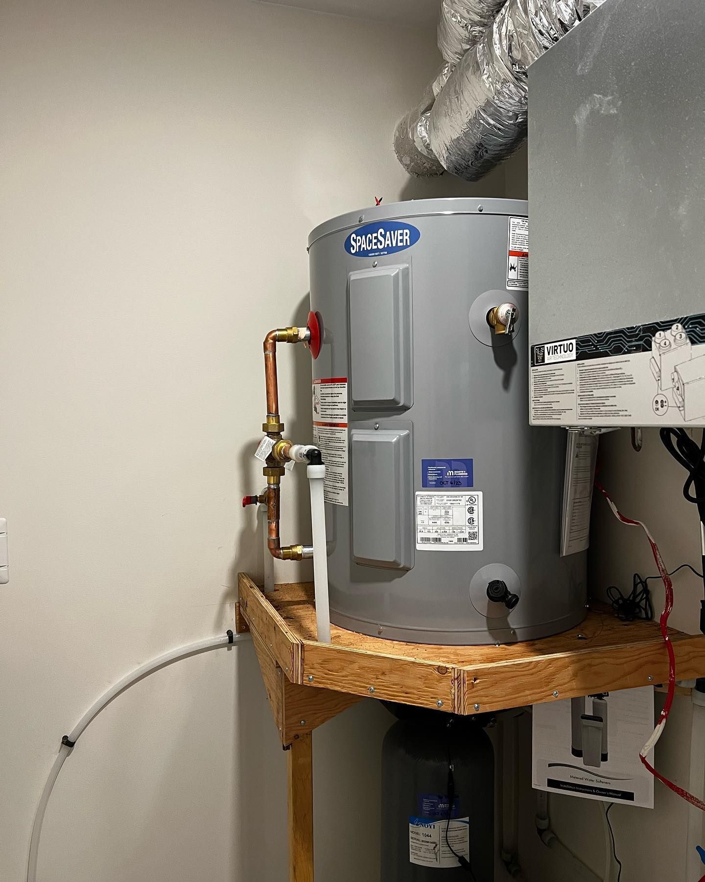 A grey electric water heater sits on a wooden platform in a utility closet with copper piping and white drain tubing.