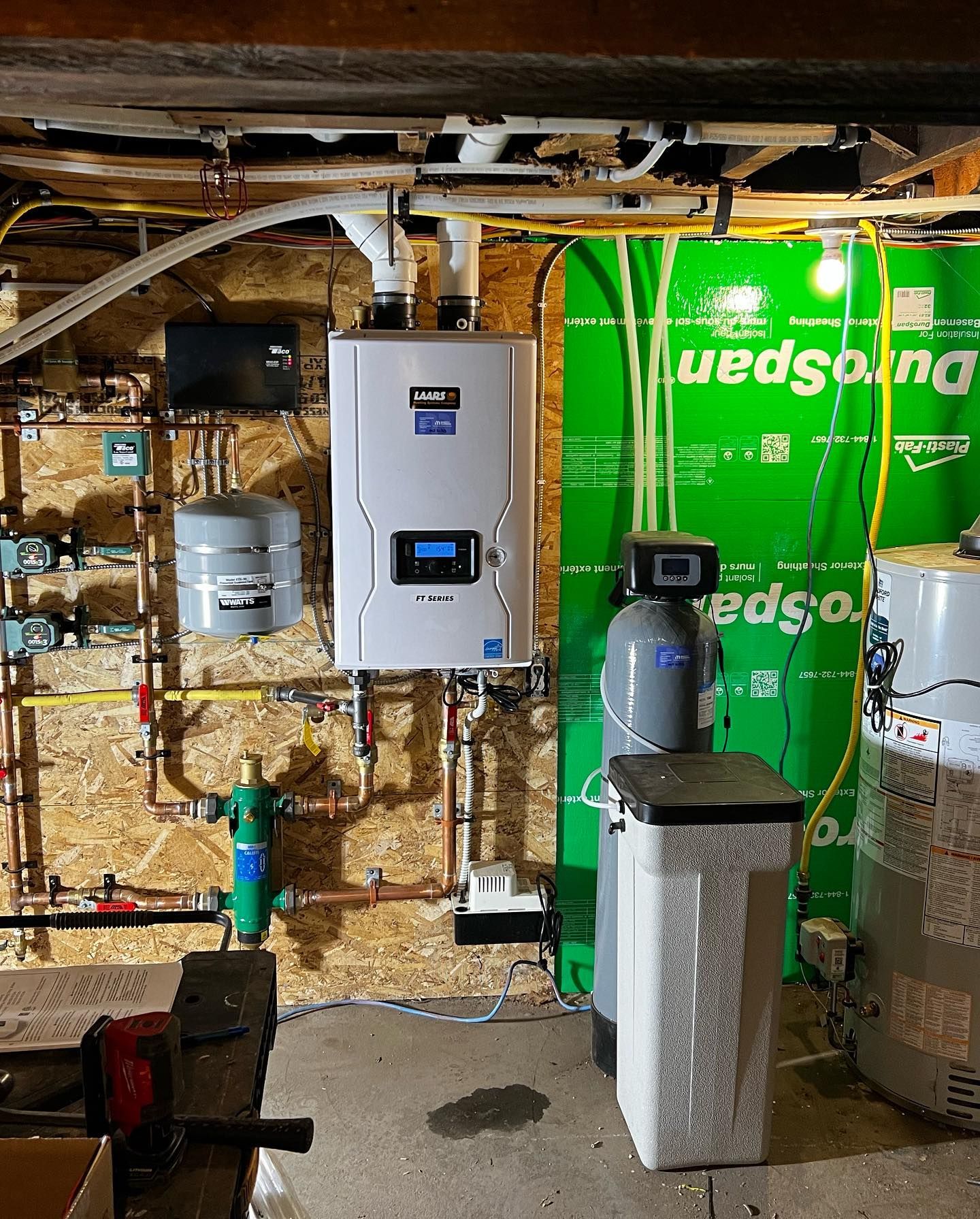 A white Navien tankless water heater, a grey water softener, and pipes mounted on a wall in a basement utility room.