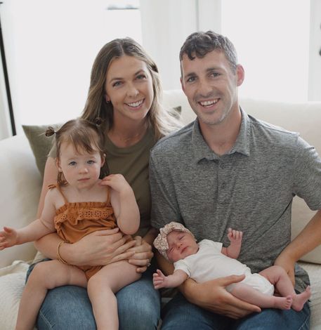 A smiling family of four sitting on a couch, holding two young children in a bright living room.