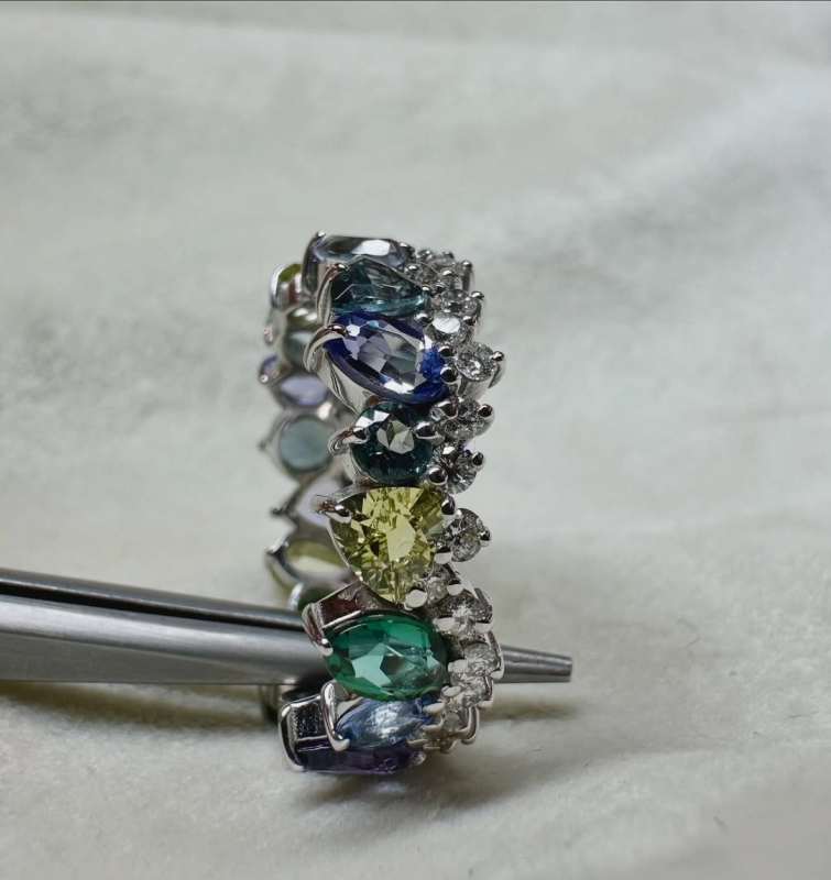 Anello in argento con gemme raggruppate, tra cui pietre blu, verdi e di colore chiaro, su una bacchetta di metallo.