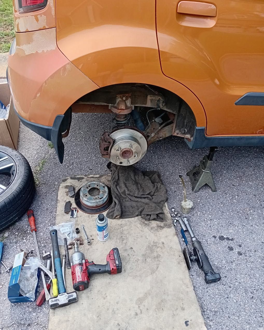 Orange car with rear wheel removed, exposing brake rotor and tools scattered on the ground during repair