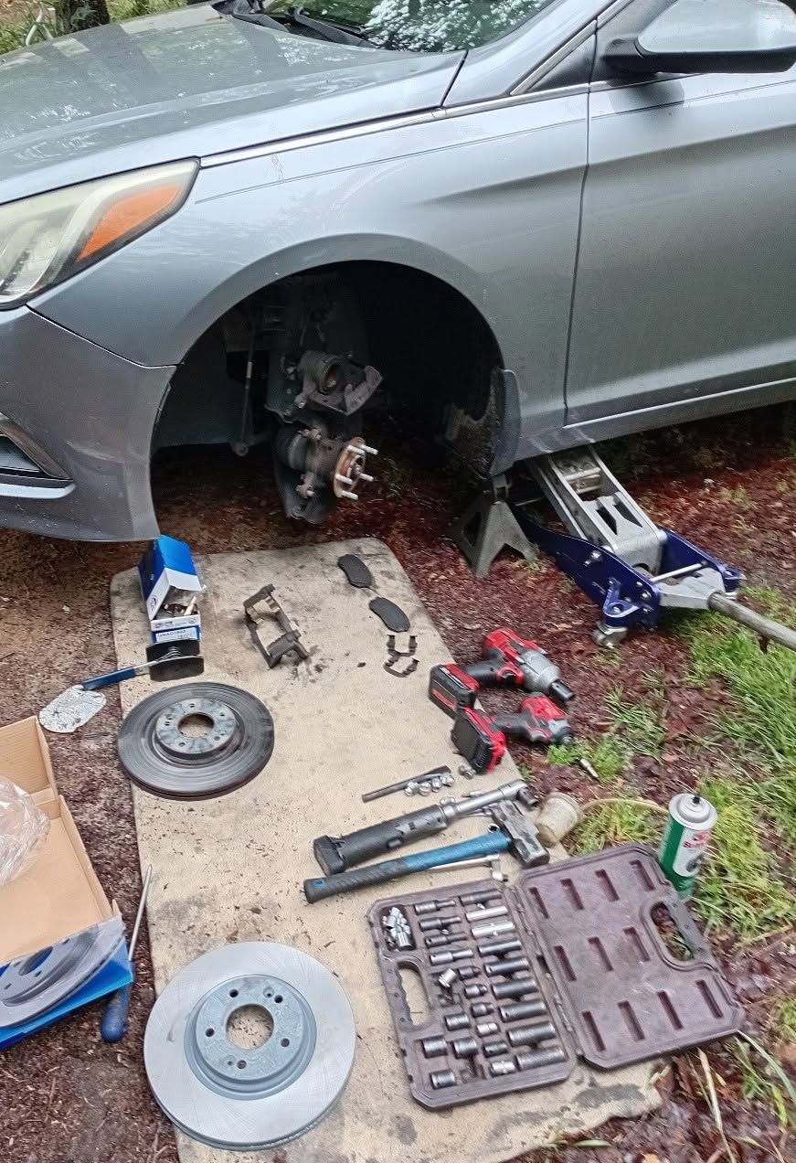 Silver car jacked up with front wheel removed, tools, parts, and brake components laid out on the ground