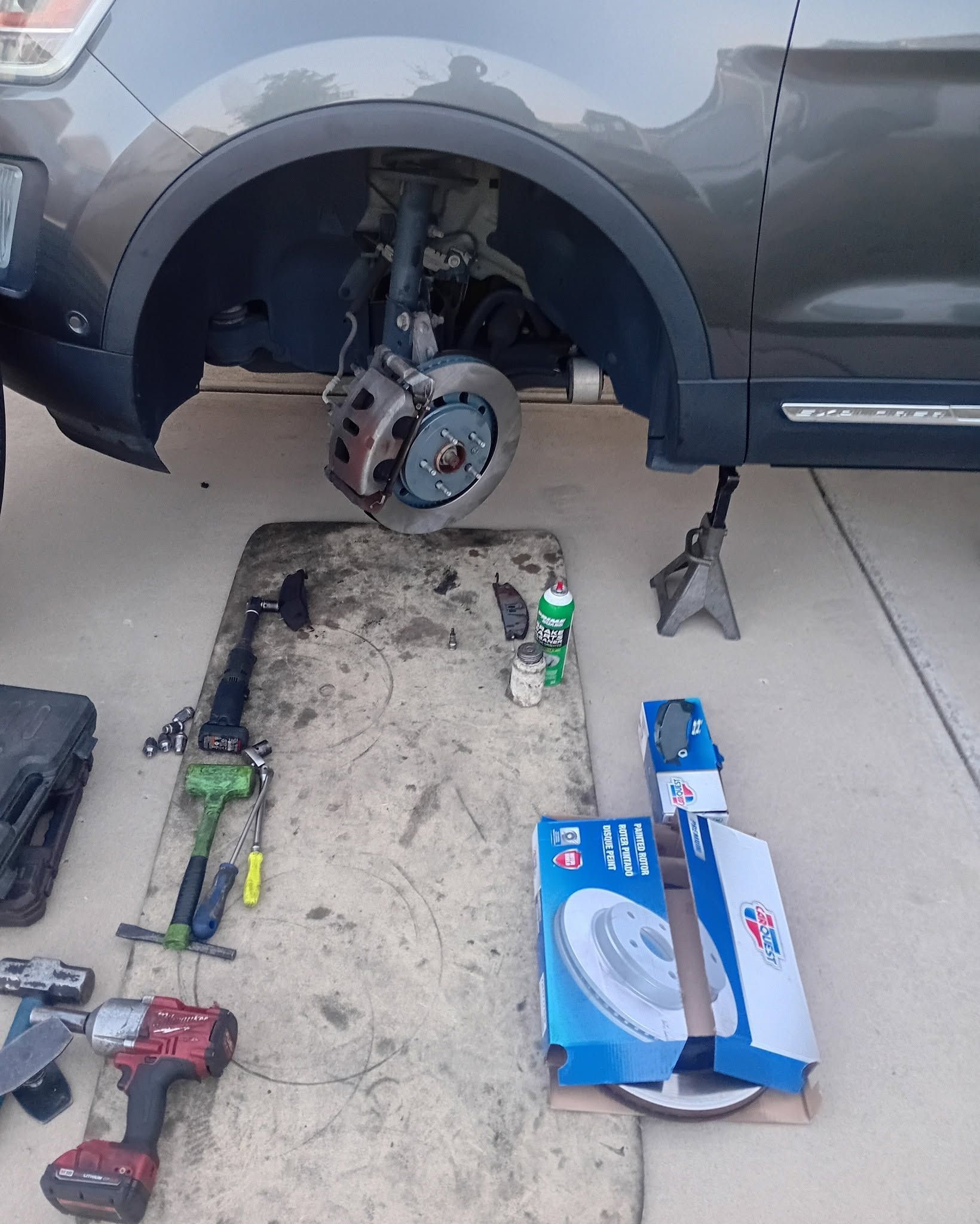 Car on jack stands with front wheel removed; tools and parts laid out on driveway.