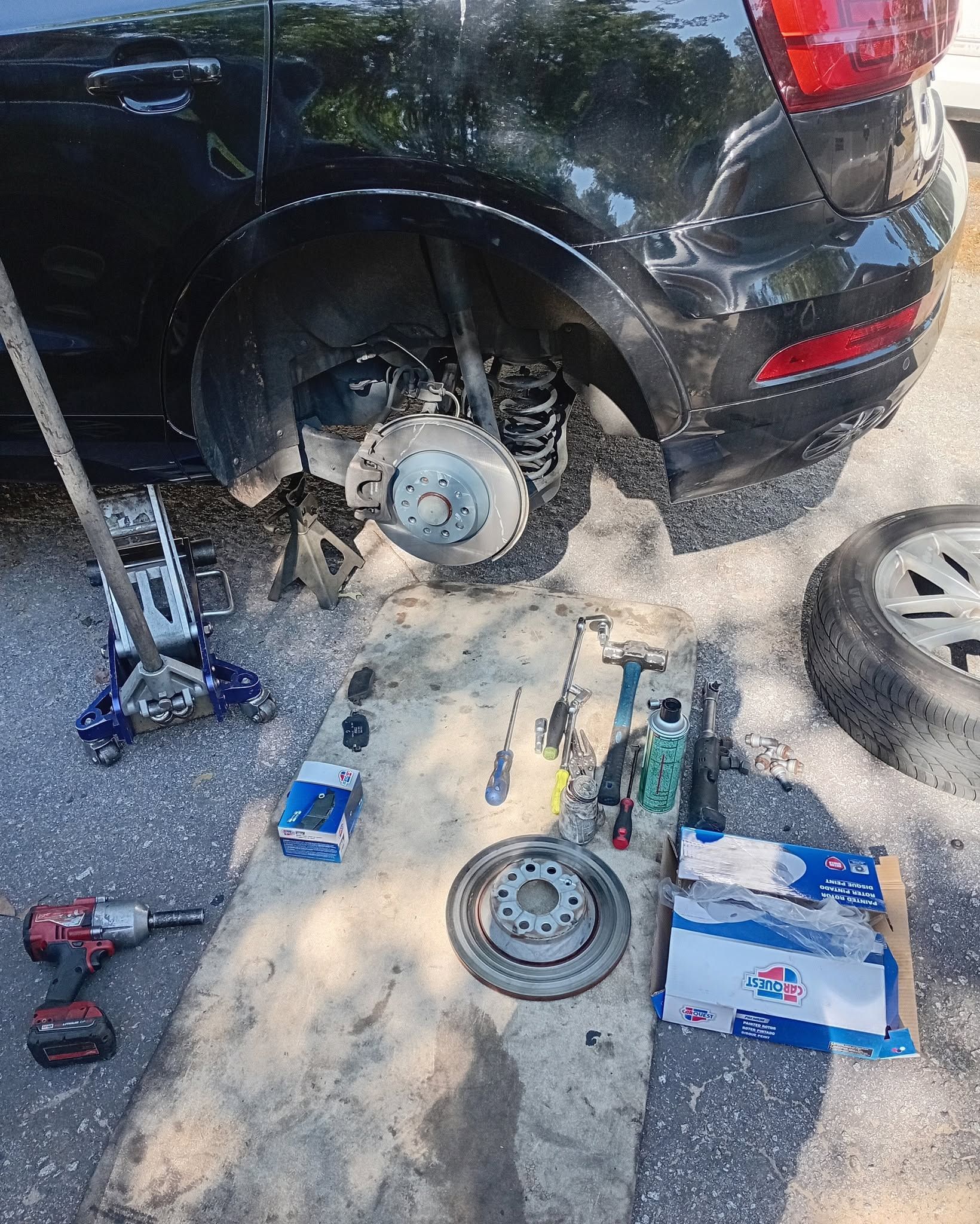 Black SUV with rear wheel removed, tools scattered on pavement, and a spare tire nearby.