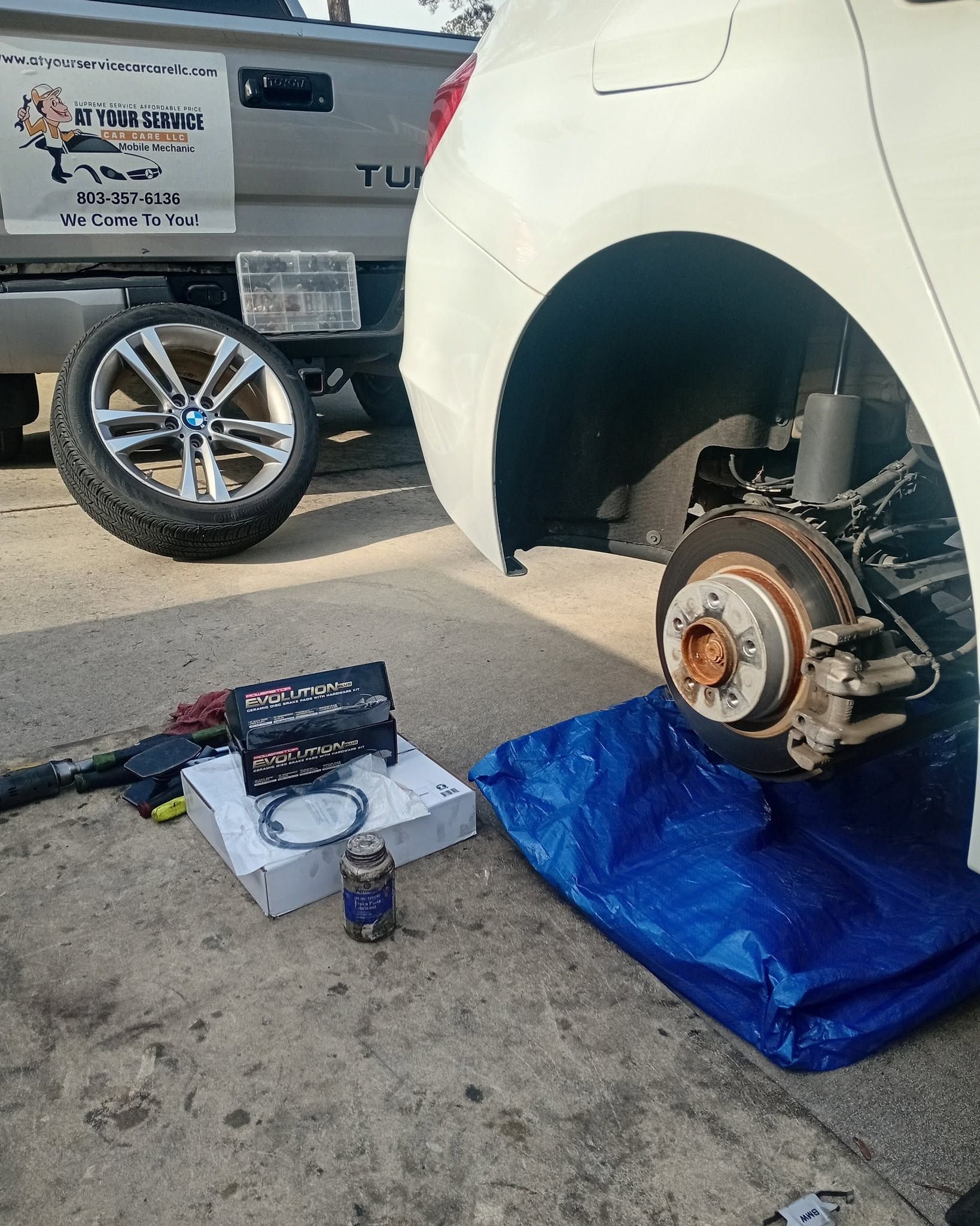Car on a blue tarp with front wheel removed, brake rotor exposed, tools and parts on the ground.