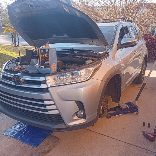 Silver SUV with hood open on a driveway, resting on a jack with tools nearby during repairs