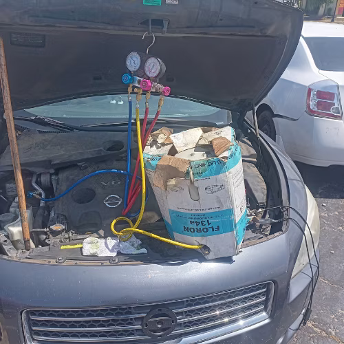 Car hood open with car engine being serviced, hoses and tools visible, bucket and vacuum equipment in front.