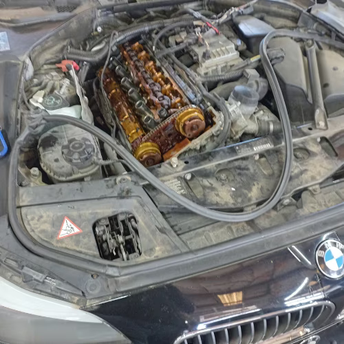 BMW engine bay with valve cover removed, exposing engine internals and hoses.