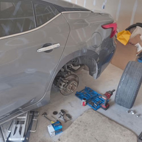 Gray car with rear wheel removed in a garage, tools and a jack on the floor, spare tire nearby