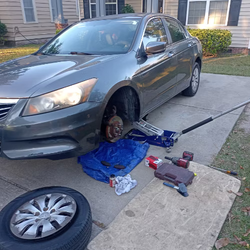 Car with front wheel removed on a driveway, tools and a spare tire nearby, mechanic working underneath