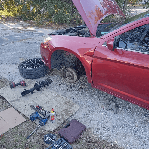 Red car with hood open and front wheel removed; tools scattered on gravel beside it.