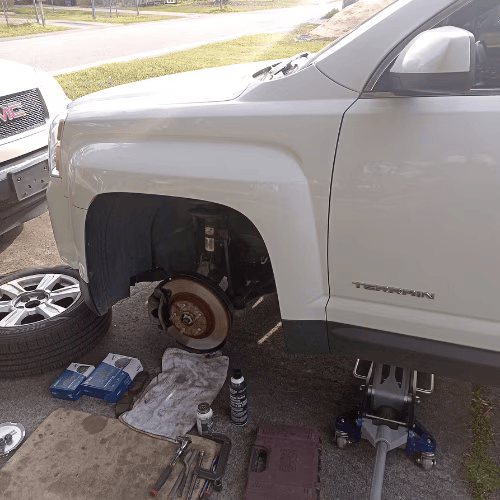 White pickup truck lifted on jack stands with front wheel removed, showing brake rotor during repair.