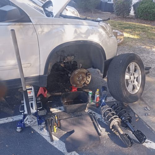 White SUV on jack stands with front wheel removed, exposing the brake assembly and suspension during repair.