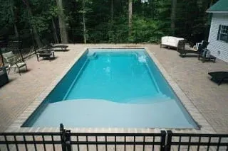 Rectangular outdoor pool surrounded by a tan deck and trees, viewed through a black fence