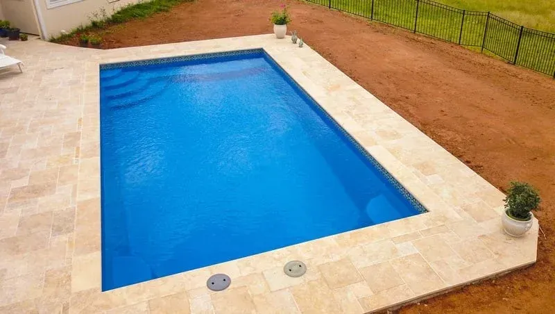 Rectangular blue swimming pool surrounded by beige stone patio and red gravel border