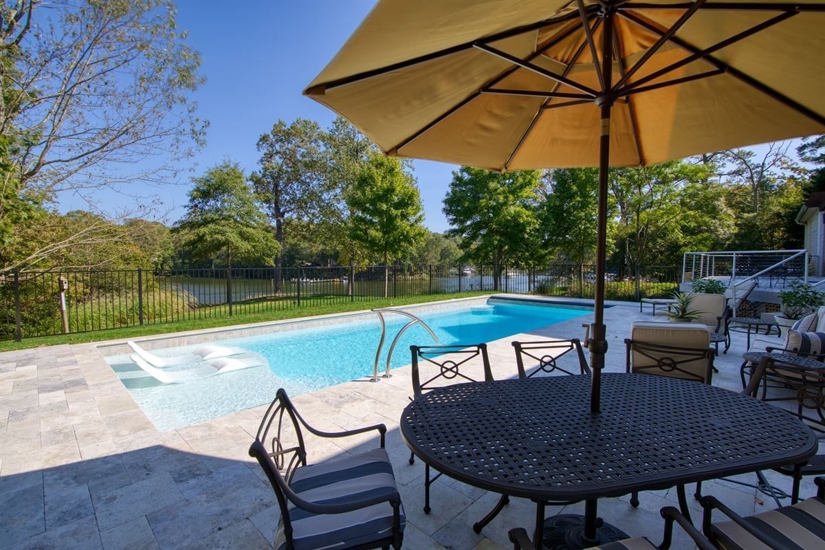 Patio with outdoor table and umbrella beside a backyard swimming pool and trees