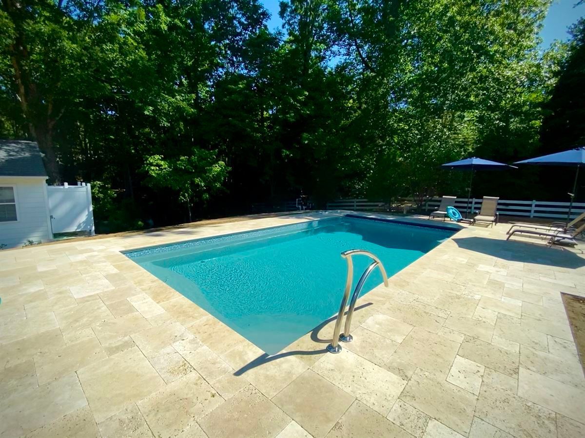 Backyard swimming pool with stone patio, surrounded by trees and lounge chairs under umbrellas
