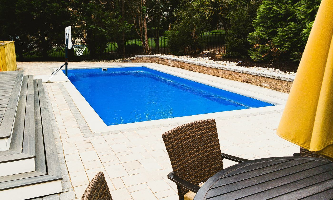 Backyard swimming pool with white deck, wicker chair, and yellow umbrella in a wooded setting