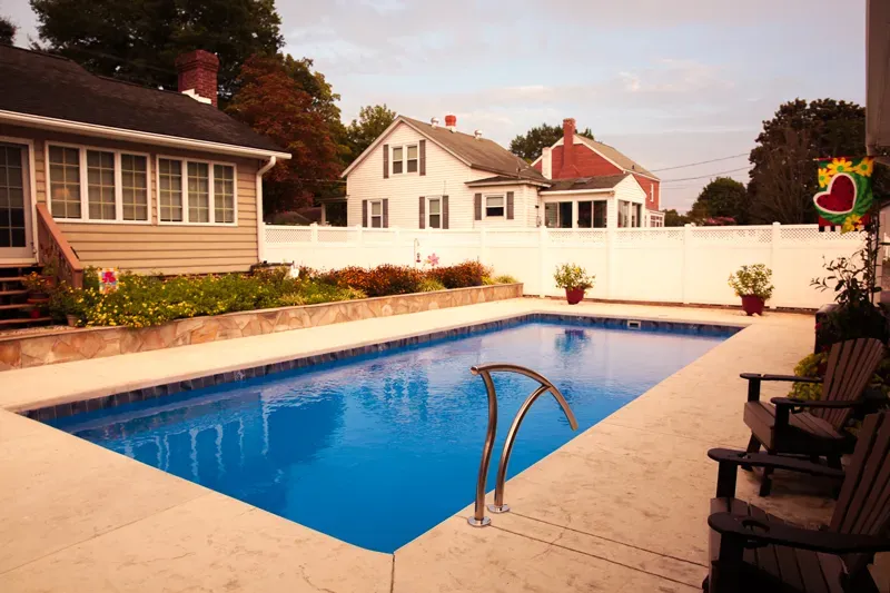Backyard swimming pool with blue water, tan patio, lounge chairs, and white fence beside houses