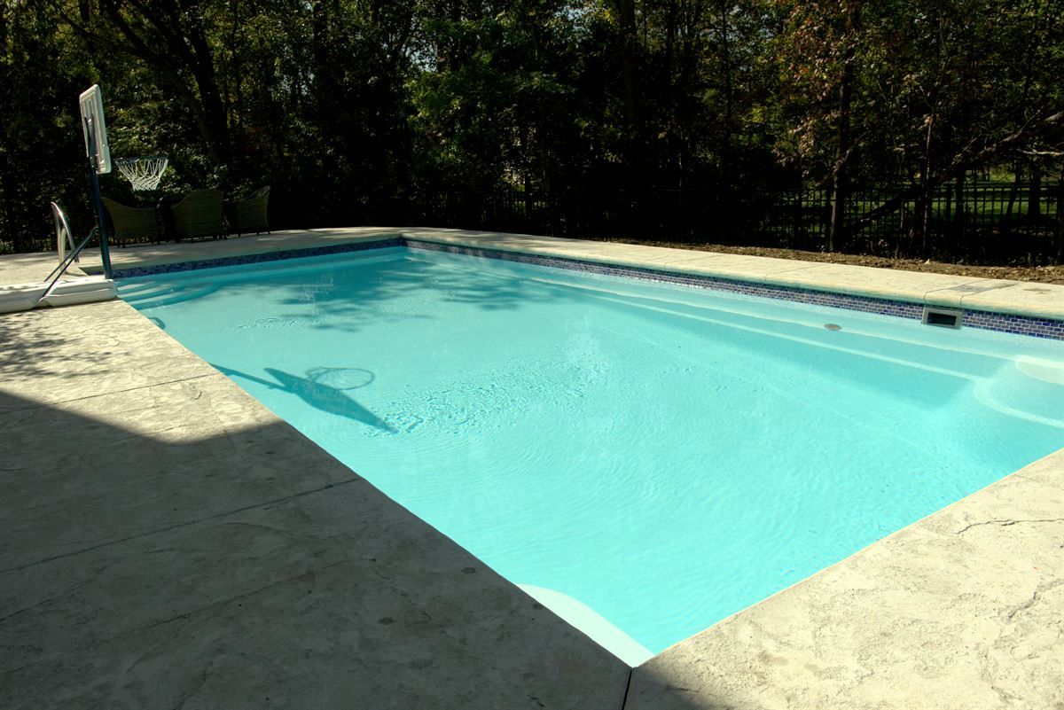 Rectangular outdoor swimming pool with clear blue water, surrounded by a light concrete deck and trees.