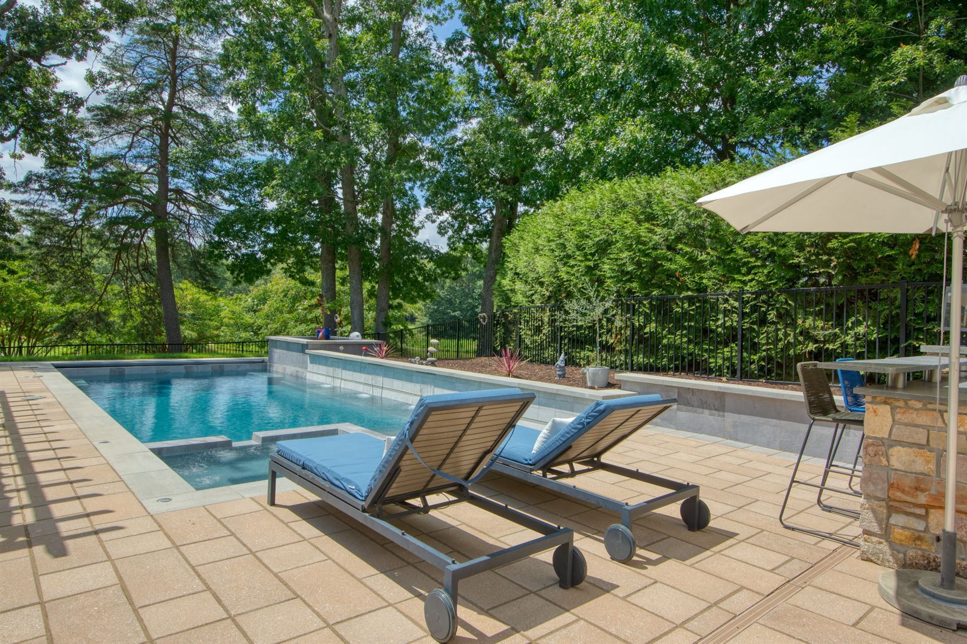 Backyard pool with two blue lounge chairs beside a stone patio and white umbrella, surrounded by trees