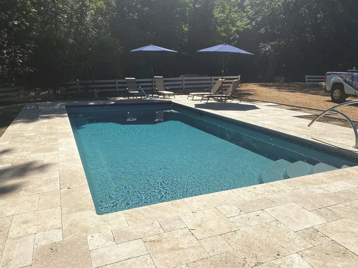 Backyard swimming pool with stone deck, lounge chairs, and two blue umbrellas in sunlight