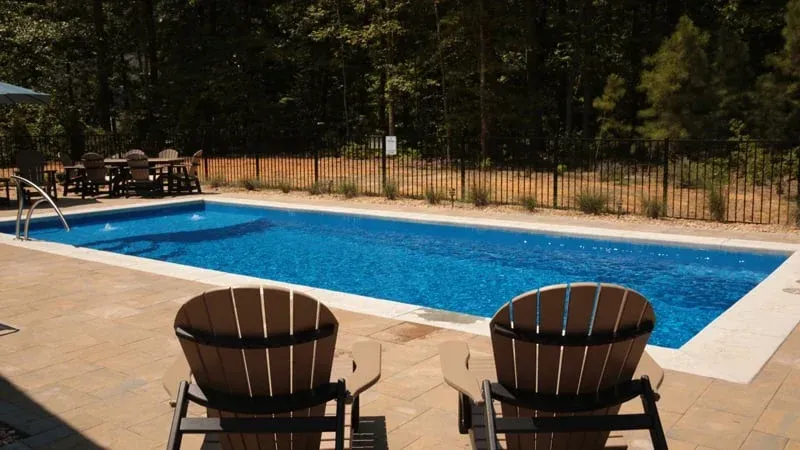 Backyard swimming pool with two brown lounge chairs on a stone patio and a fence around it