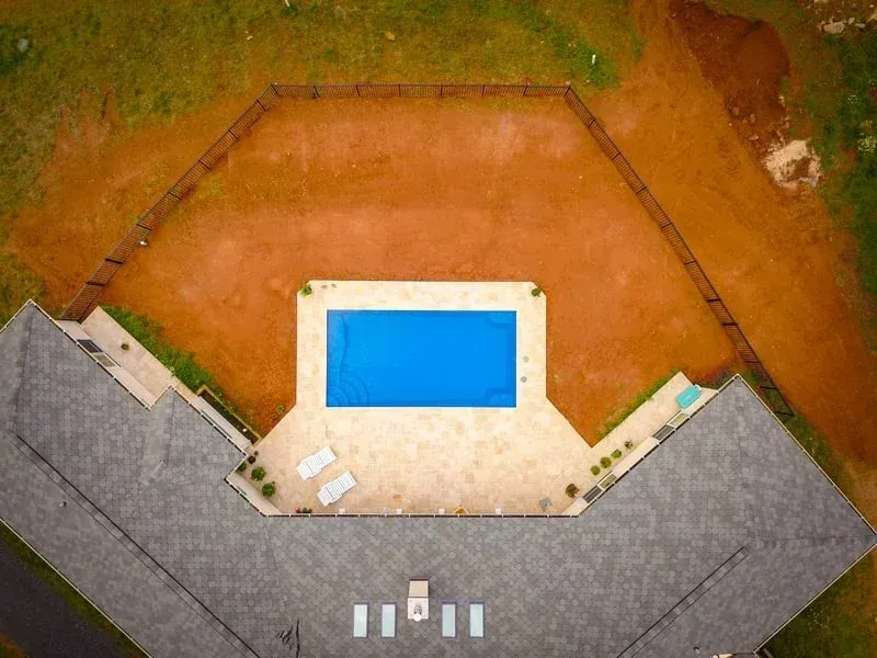 Aerial view of a backyard with a blue rectangular pool, patio, and surrounding grass and pavement.