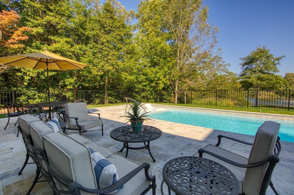 Backyard patio with cushioned seating, umbrella, and a swimming pool surrounded by trees