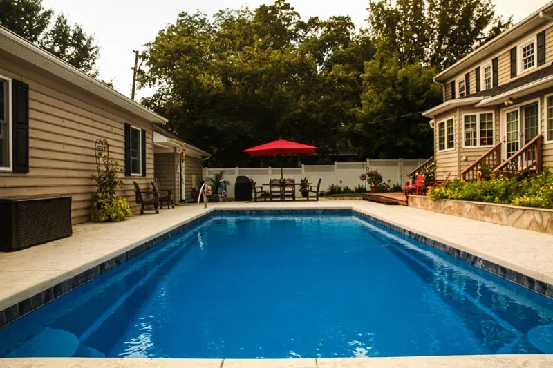 Backyard swimming pool between two houses, with a red patio umbrella and chairs at the far end.