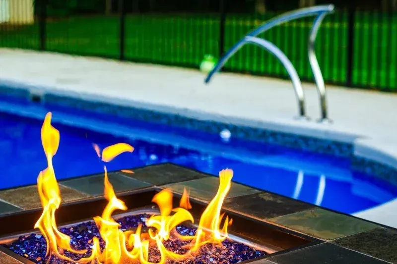 Flames from a gas fire pit beside a blue swimming pool with metal handrails