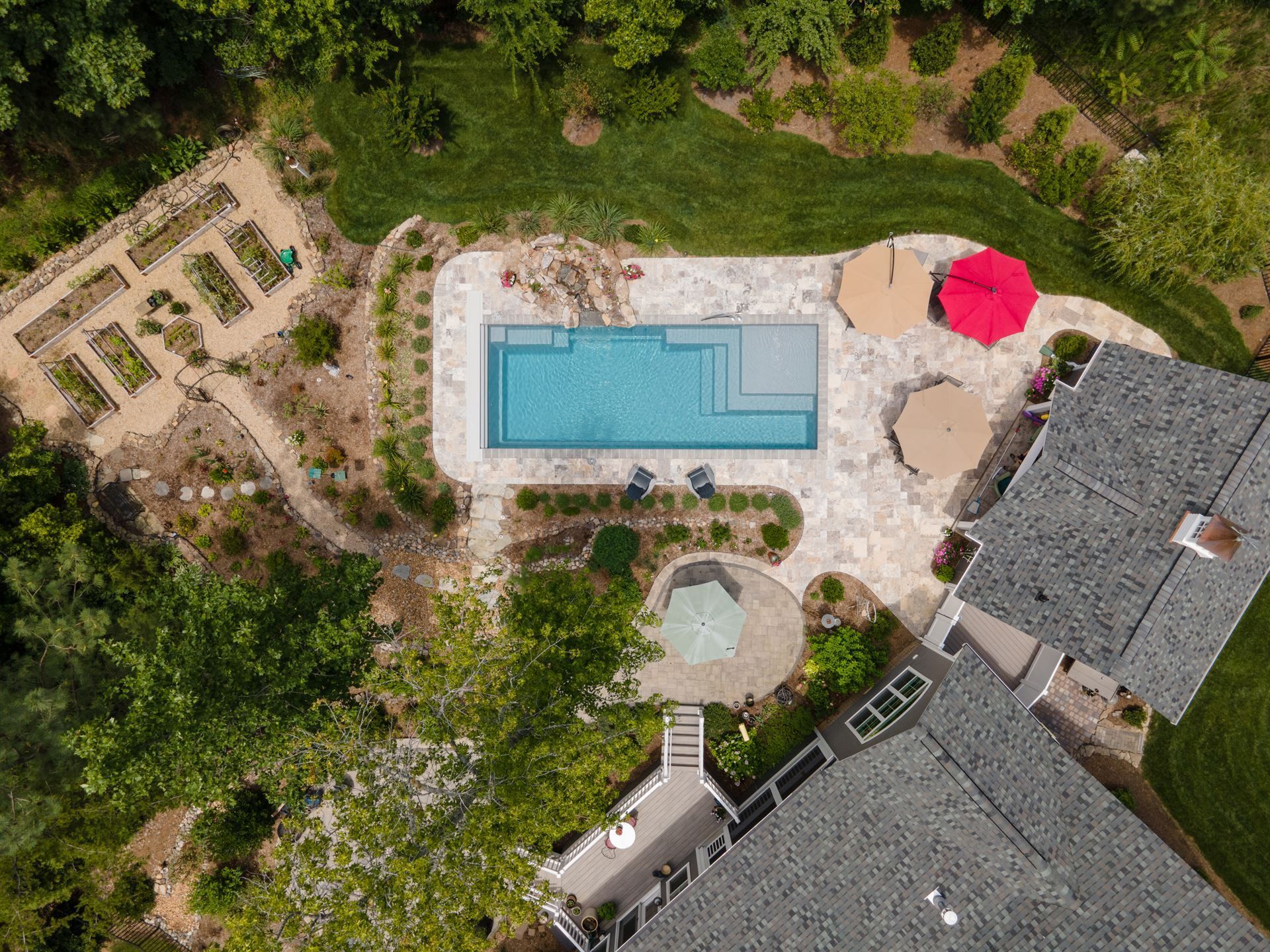 Aerial view of a backyard with a rectangular pool, patio seating, and surrounding garden beside a house