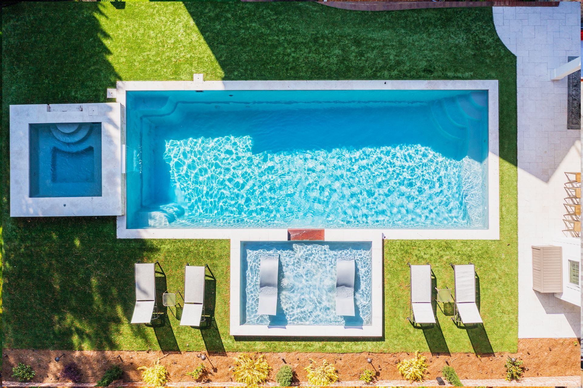 Aerial view of a turquoise pool with lounge chairs, patio seating, and a small hot tub surrounded by grass.